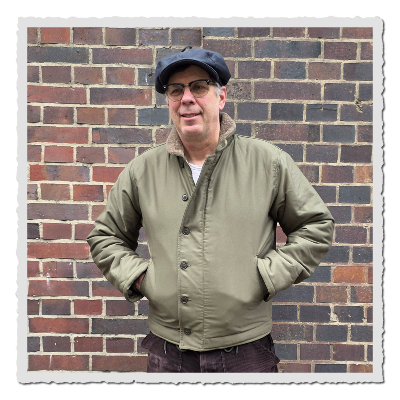 Mann vor Ziegelmauer lächelt in der grünen 1943 N1 Deck Jacket Bangor Green mit Wollkragen.